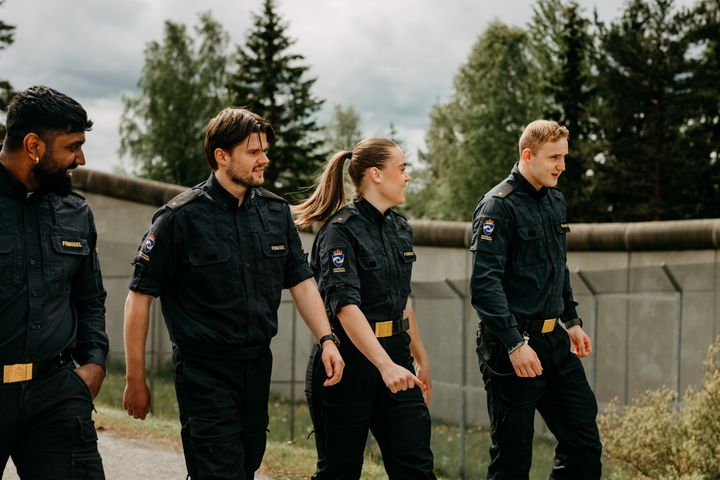 Fire personer i kriminalomsorgens uniform går langs en mur i et utendørsmiljø.