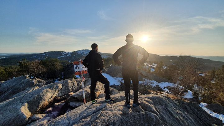 To menn står med med armene i siden, med ryggen vendt mot kamera og speider mot sola på toppen av et fjell. T