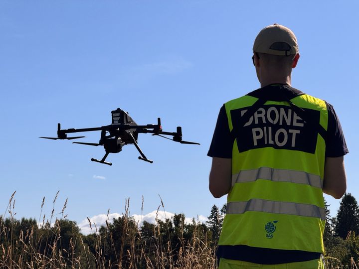 Ryggen til en mann, blå himmel og drone i lufta foran han over toppen av skogen i horisonten. På mannens rygg er det en refleksvest hvor det står "dronepilot"