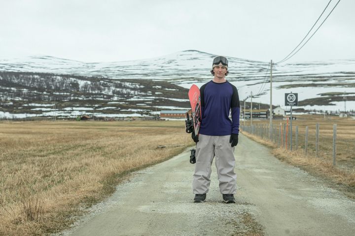 Mann i snowboardutstyr står på en grusvei i et fjellandskap med snødekte topper.