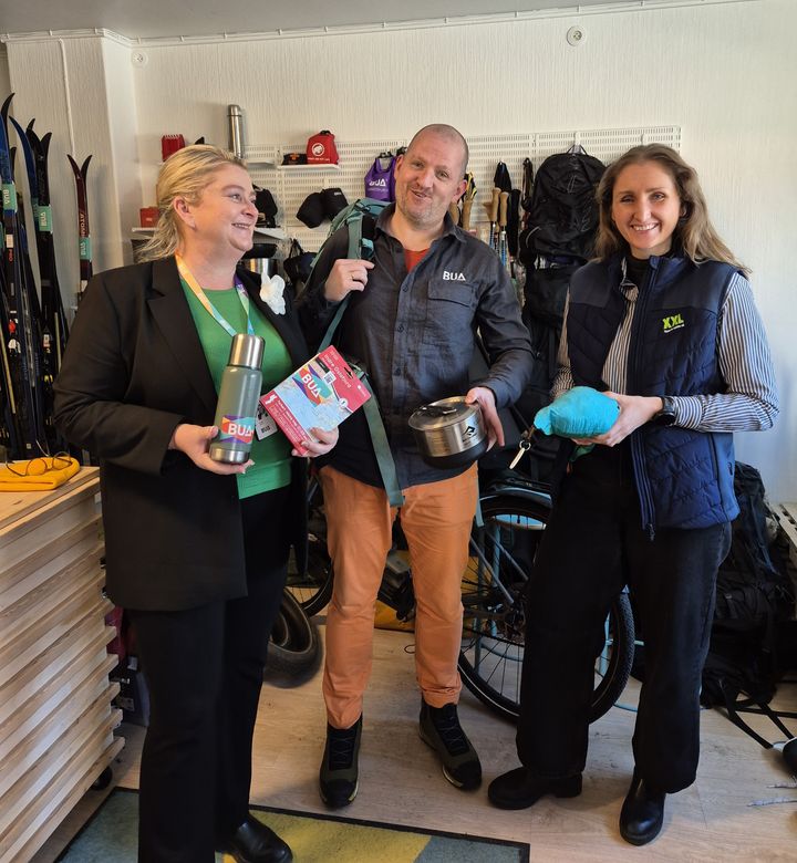 Kort tekstbeskrivelse: Tre personer står sammen innendørs og holder ulike produkter. Bak dem vises sportsutstyr som ski og sykler.