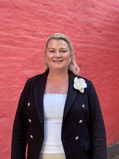 For a bildet: En kvinne står foran en rød vegg. Hun har på seg en sort jakke med en hvit blomst detalj.