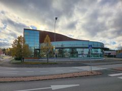 Alt-tekst: En rund bygning med glassfasade i et handelsområde, med trær i høstfarger og en vei med fotgjengerfelt i forgrunnen.