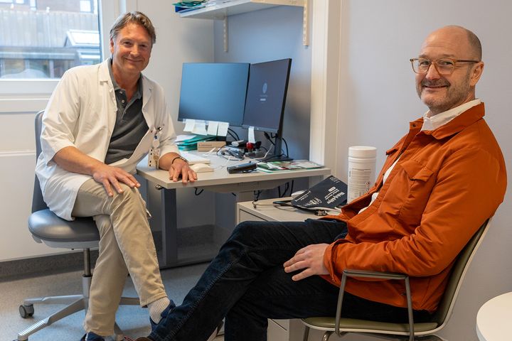 Mannlig%20kreftlege%20med%20hvit%20sykehusfrakk%20og%20voksen%20mann%20med%20briller%20og%20oransje%20jakke%20sitter%20mot%20hverandre%20p%E5%20et%20legekontor.