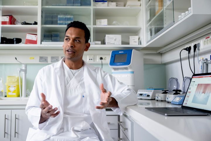 Mann lege i hvit frakk gestikulerer  sittende i et laboratorium