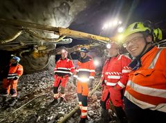 Bildet er tatt inne i tunnelen med en gul fjellboremaskin i brakgrunnen og i skinnet fra hodelykert og maskinens arbeidslys. Bildetektsen sier, Skanska passerer 15 000 meter drevet tunnel på OPS Hålogalandsvegen.  I marmorhvelvet i Gullberg øst, som vender ut mot Tjeldsundbroen står fra venstre: Statens vegvesen ved Bjørn-Einar Hansen, Hålogalandsvegen AS ved Kent Gøran Johansen, Statens vegvesen ved Henning Larsen og sprengningsbas Tore Gunnar Gundersen.