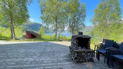 Bildet viser en stor platt ute bak vertshuset med en utepeis i naturstei, høye bjørker med nye grønne blader og utsikt over en speilblank fjord. Bildeteskten sier, Illsted med mening: Idyllisk uteplass med utepeis i naturstein og herlig utsikt over Sørfjorden.