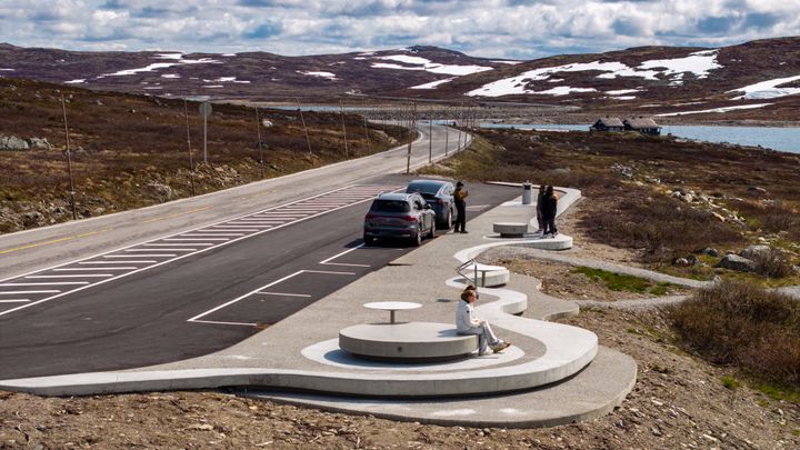 To ungjenter sit på runde rastemøblar i betong og ser på utsikta over innsjøen Ørteren på Hardangervidda. Nokre turistar tek bilete av kvarandre i bakgrunnen.