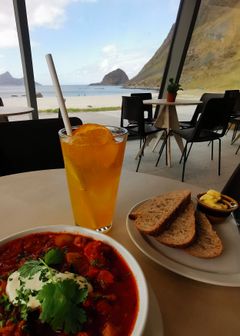 Bilde viser et bord med appelsinjuice, brød og smør og en tallerken med suppe. Utsikt til strand og fjell i bakgrunnen.