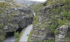 Bildet er et dronebilde av E45 der vegen slynger seg langs Eibyelva igjennom en canyon med stupbratte fjell på begge sider. Bildeteksten sier, Flaskehalsen: Dagens vei gjennom kløfta følger en naturlig elvecanyon som skjærer seg ned igjennom fjellet fra Finnmarksvidda mot Alta. Fjellet har dårlig kvalitet og det går ofte snøskred, sørpeskred og steinras på strekningen.