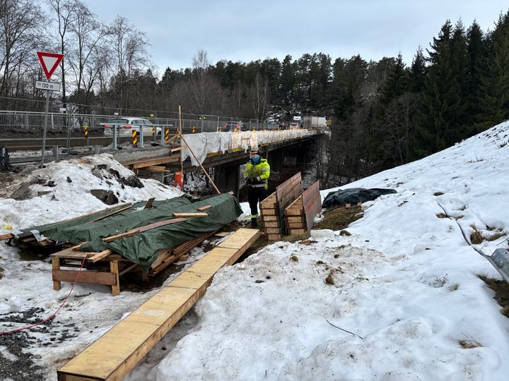 Bru under rehablitering i bakgrunnen, materialer og en arbeider i forgrunnen står ved siden av brua.