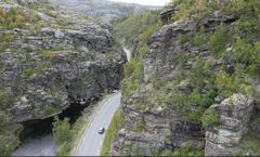 Bildet er et dronebilde som viser E45 dypt nede i en evledal. Bildeteksten sier: Flaskehalsen: Dagens E45 gjennom kløfta følger en naturlig elvecanyon. Den skjærer seg dypt ned i fjellet fra Finnmarksvidda mot Alta. Fjellet er dårlig og porøst. Snøskred, sørpeskred og steinras gir ofte stengt veg. Nå skal strekningen legges i tunnel.