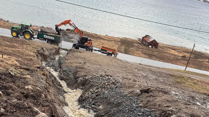 Bildet viser en bekk med stor vannføring og to anleggsmaskiner der bekken møter dagens E10. Bildeteksten sier: Må stenge vegen: En bekk ved Hårvik i Tjeldsund må sikres og legges i rør under dagens E10 for å hindre utgraving av veibanen. Jobben skal gjøres på sein kveld og natt.