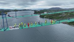 Bilete av første delkabel midtfjords. Arbeidarear frå NRS "føljer" kabelen over fjorden via catwalken. Delkabelen (til høgre i bilete, med flagg for anledninga) vert trekt over fjorden. Tilsaman 82 slike delkablar vert innstalert. Arbeidet er venta å ta omlag tre månder.