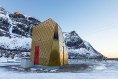 En toalettbygning i gyldne farger og med rød dør står i et kystlandskap omgitt av spisse, delvis snødekte fjell.