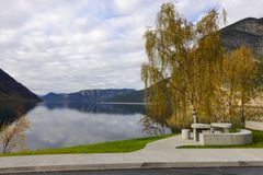 Betongbenkar og bord ved Lærdalsfjorden på Røyrnes. I bakgrunnen ligg fjorden omgjeven av fjell og haustfarga bjørketre.