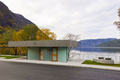 Toalettbygg i betong og glas på Røyrnes ved Lærdalsfjorden. Framfor bygget ligg asfalt og gangareal, medan fjorden, fjell og haustfarga tre dannar bakgrunnen.