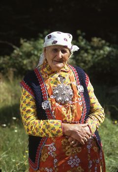 Bilde av en gammel kvinne med fargerike klær og stor sølje på brystet.