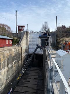 Sluseanlegg i kanal, med mannskap i arbeid, sluseporten godt synlig, røde driftsbyninger langs kanalen og blå himmel i bakgrunnen.