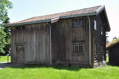 På en gressplen står et forvitret trehus med tegltak, små vinduer og en dør på siden.