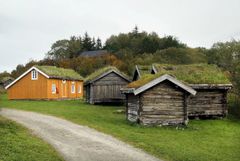 En grusvei går mellom et gult hus og gamle treboder, alle med gresskledde tak, som ligger på en gresslette med trær i bakgrunnen.