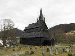 En stavkirke i tre med bratt tak og høyt spir står på en kirkegård, omgitt av gravsteiner og åser under en overskyet himmel.