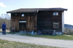 En person står ved en gammel trebygning med tegltak, benker og spredte utendørsgjenstander på et gressområde ved siden av en grussti.