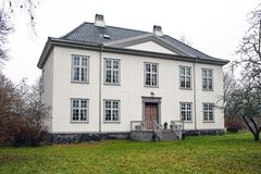 Et toetasjes trehus med hvit kledning, store vinduer og grått tegltak, plassert på en gresskledd hage med nakne trær.