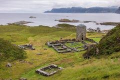 Gamle steinruiner og et tårn står på gresskledde klipper med utsikt over havet, med små øyer og fjell i det fjerne under en overskyet himmel.