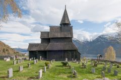 En stavkirke i tre med et høyt tårn i midten står på en kirkegård, omgitt av gravsteiner, med fjell og en innsjø i bakgrunnen.