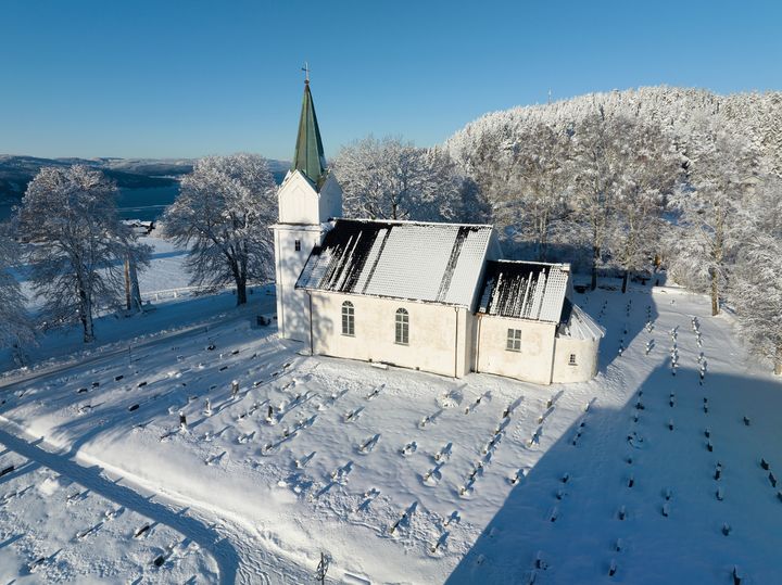 Flyfoto av en hvit kirke med et grønt kirketårn omgitt av en snødekt kirkegård og trær på en klar vinterdag.
