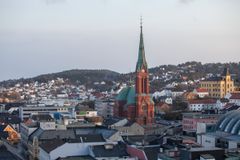 Et bybilde med en fremtredende kirke i rød teglstein med et høyt, grønt spir, omgitt av bygninger og hus på bølgende åser under en klar himmel.