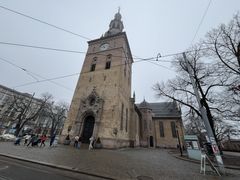 En stor steinkirke med klokketårn står på en brosteinsbelagt gate; folk spaserer i nærheten, og løvløse trær omkranser bygningen på en overskyet dag.