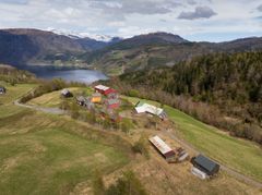 En liten klynge gårdsbygninger ligger i en gressbevokst åsside med en innsjø, skog og snødekte fjell i bakgrunnen under en delvis overskyet himmel.