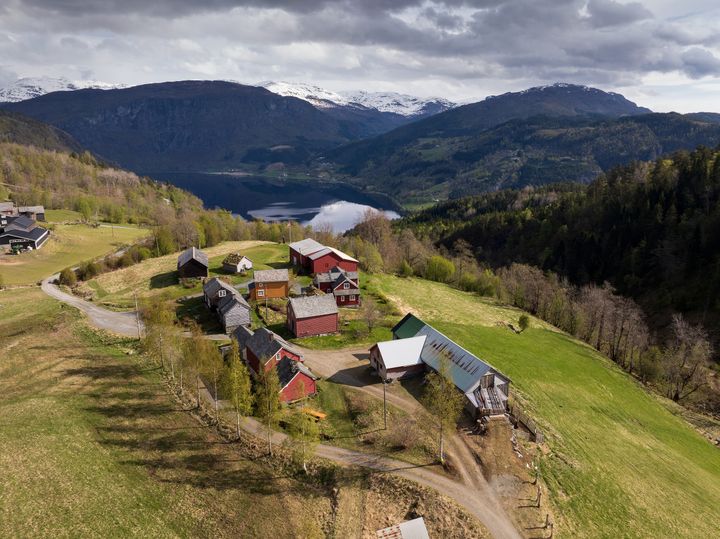 Flyfoto av en liten landsby med røde og hvite bygninger, omgitt av grønne åser, trær og fjell i det fjerne, nær en innsjø under en overskyet himmel.