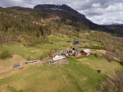 Flyfoto av en gård med røde og hvite bygninger omgitt av grønne åkrer, skogkledde åser og et fjell i bakgrunnen under en overskyet himmel.