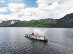 En dampdrevet båt med røyk fra skorsteinen seiler på en innsjø, omgitt av grønne åser og fjell under en delvis overskyet himmel.