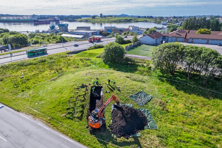 Luftfoto av et utgravningsområde ved en vei, med arbeidere og en oransje gravemaskin som graver rektangulære grøfter på et gressområde, med bygninger og vann i bakgrunnen.