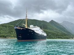 Et stort svart-hvitt skip med navnet Salten ligger for anker i rolig turkis vann nær skogkledde fjell under en overskyet himmel.