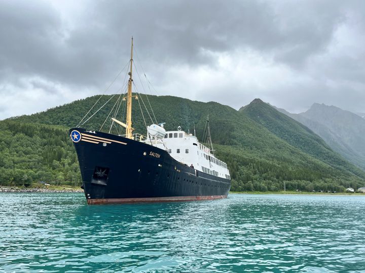 Et stort svart-hvitt skip med navnet Salten ligger for anker i rolig turkis vann nær skogkledde fjell under en overskyet himmel.