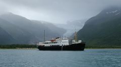Et svart-hvitt skip flyter på rolig vann, omgitt av grønne fjell og en isbre i bakgrunnen under en overskyet himmel.