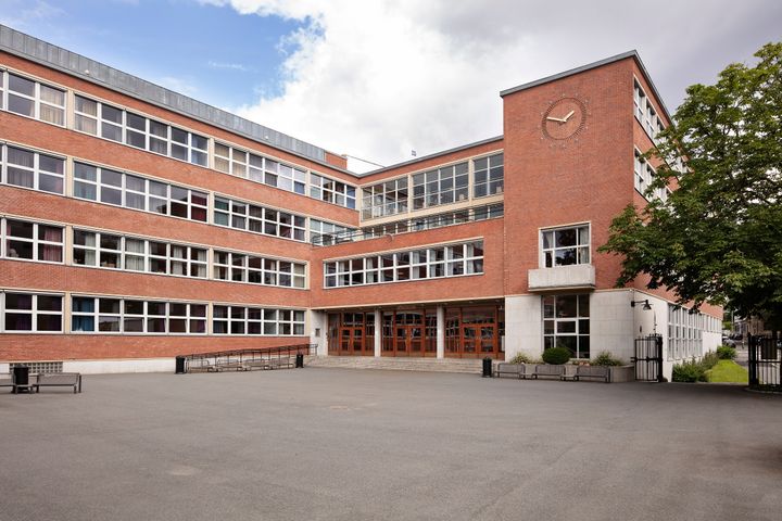 En skolebygning i rød murstein med store vinduer, et klokketårn i hjørnet og en bred, asfaltert gårdsplass ved hovedinngangen.