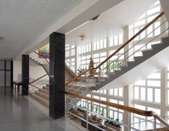 En bred trapp med steintrinn og rekkverk i tre i et solfylt atrium, med en statue av en kvinne og potteplanter.