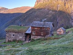 Et tradisjonelt trehus med gresstak ligger i en grønn åsside, med et lite skur i nærheten og fjellene i bakgrunnen.