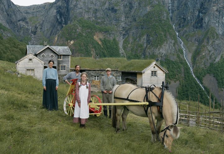 En familie står på en gresslette med en hest som trekker en gul vogn, med en rustikk gård i tre og fjell i bakgrunnen.