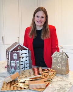 Blid kvinne med halvlangt hår og rød blazer smiler mot kamera. Hun stør ved kjøkkenbenken med ulike julekalendere foran seg.