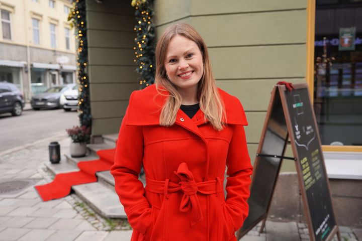 Kvinne med julerød kåpe og halvlangt hår i julepyntet bybilde smiler mot kameraet.