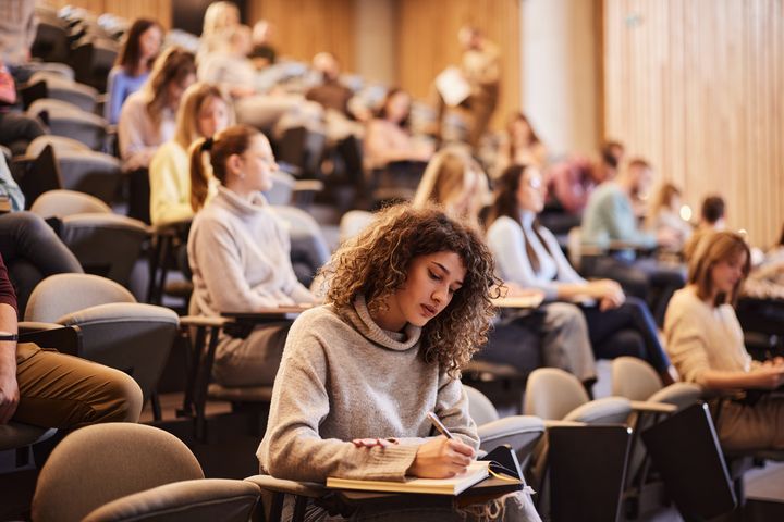 En kvinnelig student sitter og skriver i et auditorium med mange medstudenter bak seg