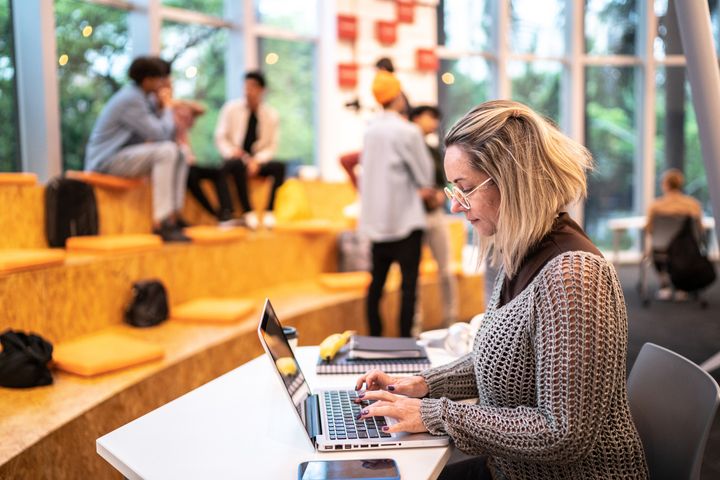 En kvinne sitter og jobber på en pc med folk som sitter på benker i bakgrunnen