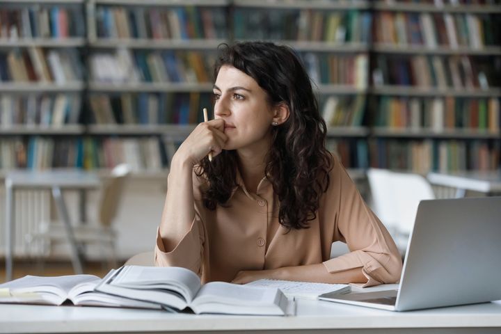 Portrettbilde av en tankefull kvinne som sitter med en pc i et bibliotek og kikker ut i lufta